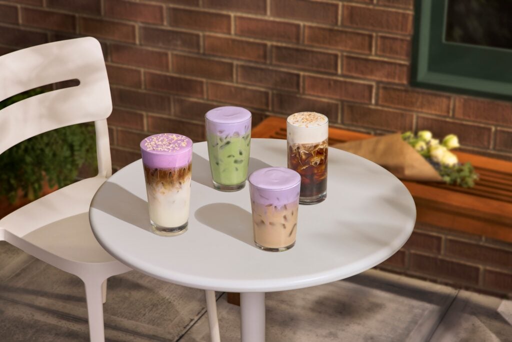 Four colorful drinks are arranged on a round white table. They include a layered iced coffee with a purple foam top, a green drink, and two other iced beverages topped with purple and white foams. A brick wall is in the background.