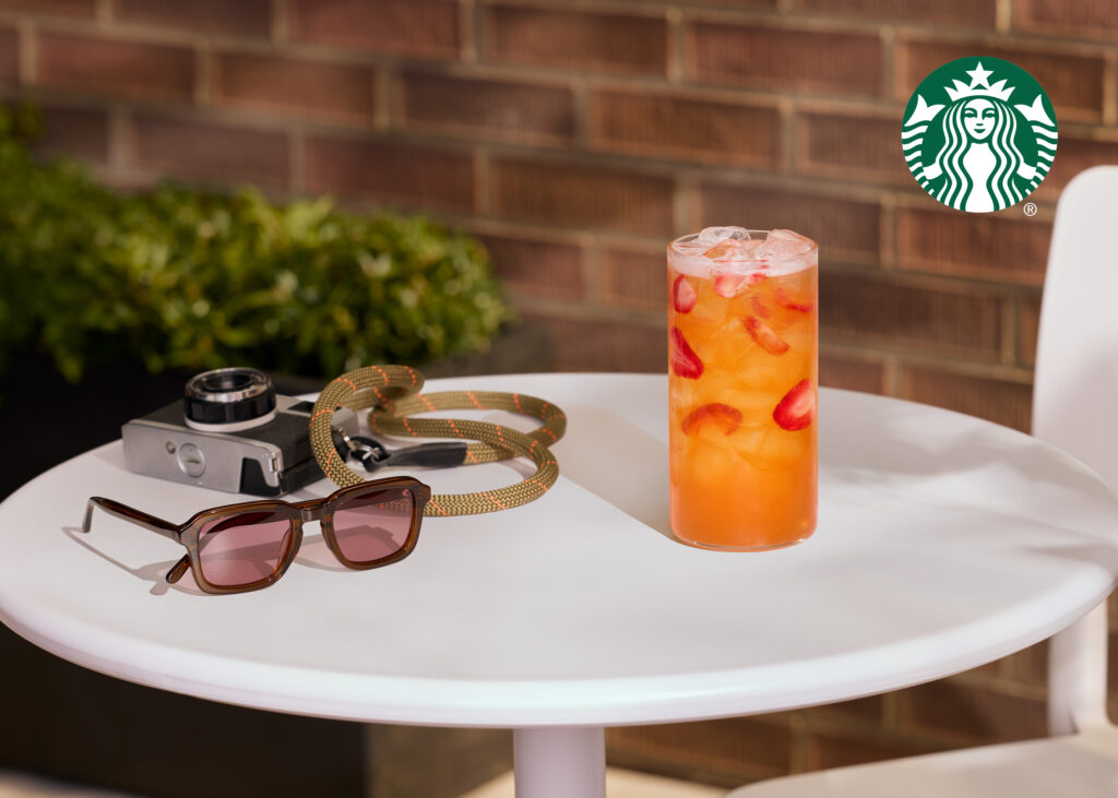 A refreshing drink with ice and strawberries sits on a white table, accompanied by sunglasses and a vintage camera, with greenery in the background.