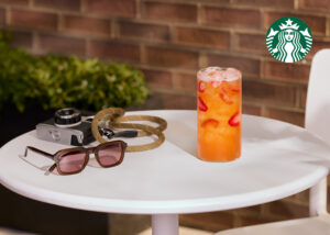 A refreshing drink with ice and strawberries sits on a white table, accompanied by sunglasses and a vintage camera, with greenery in the background.