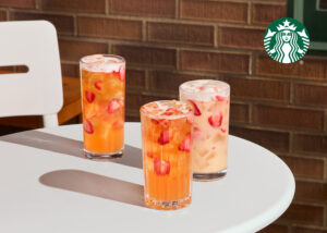 Three tall glasses of iced drinks sit on a white table. The drinks feature slices of strawberries and are in vibrant colors, with one orange, one pink, and one lighter shade. A brick wall serves as the backdrop.