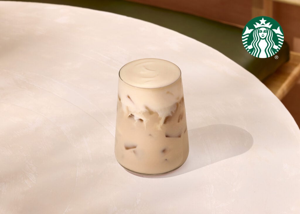 A clear glass filled with iced coffee, topped with creamy foam, sits on a light-colored table.