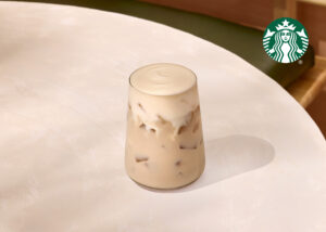 A clear glass filled with iced coffee, topped with creamy foam, sits on a light-colored table.