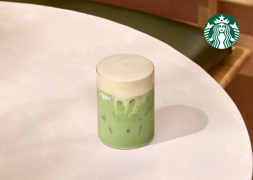 A tall glass filled with a layered green drink, topped with a frothy cream. The drink is served over ice on a light-colored table.