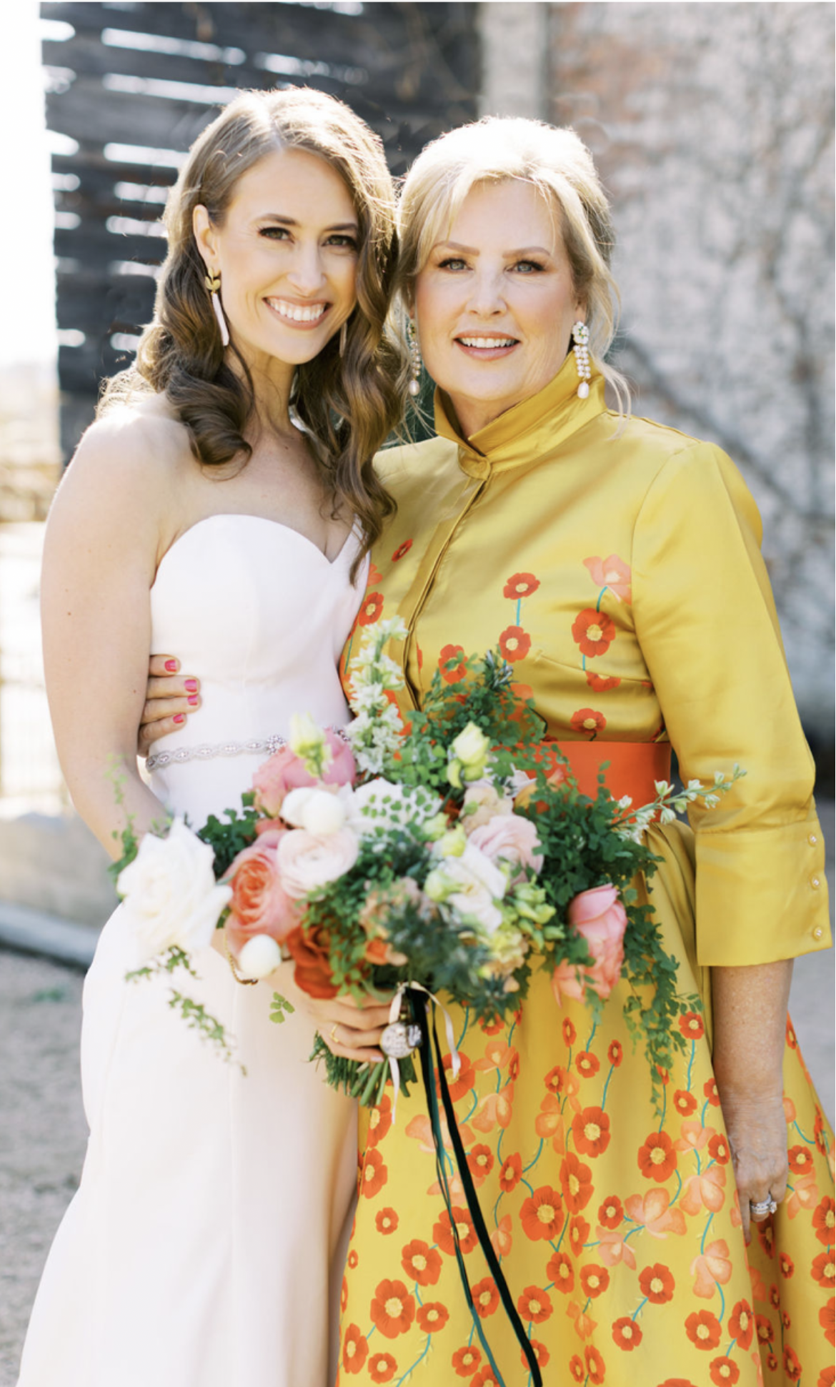 A bride in a strapless white gown smiles alongside a woman in a vibrant yellow dress adorned with floral patterns. The bride holds a bouquet of pink and white flowers.