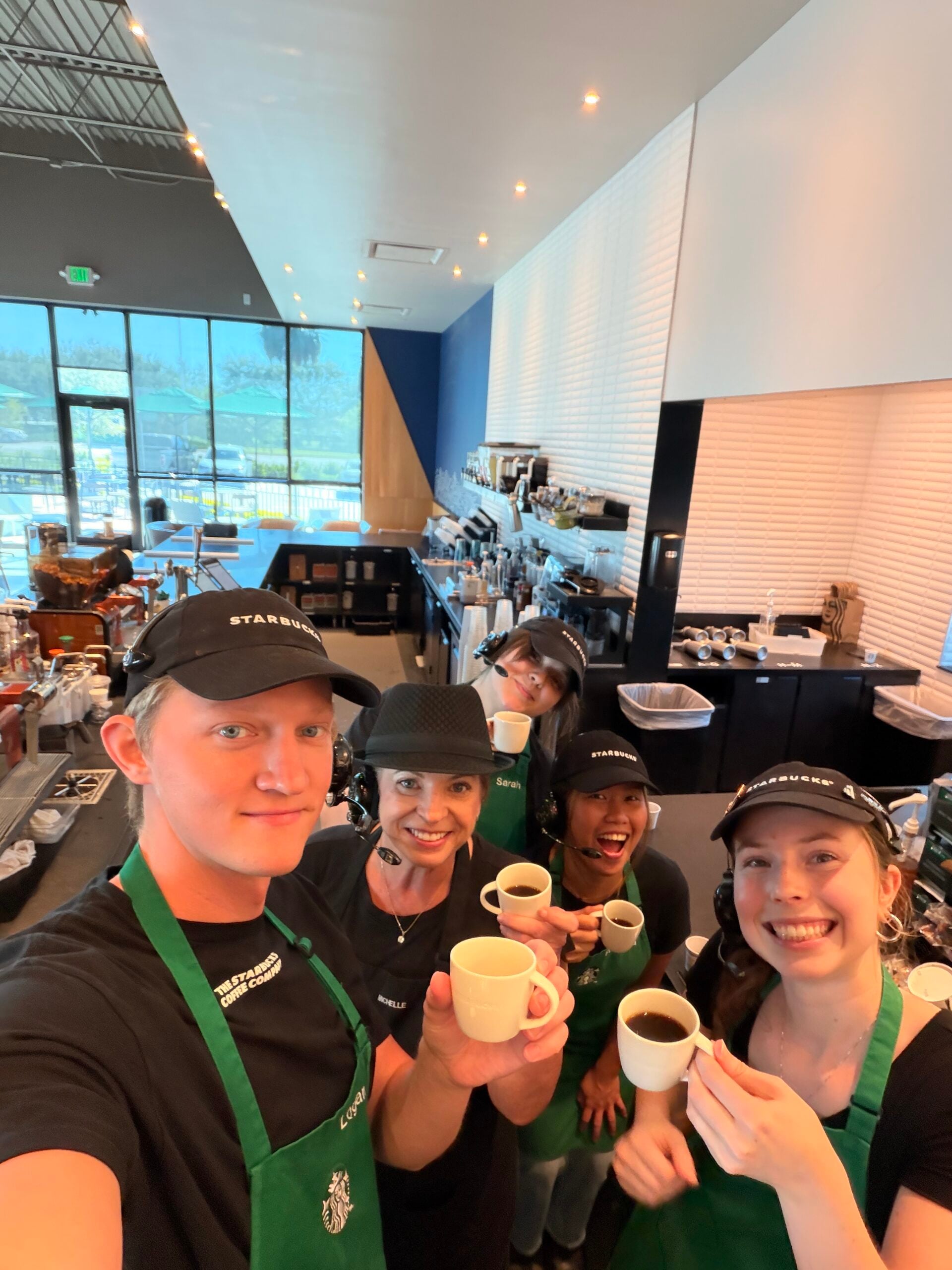 A group of five baristas in a coffee shop are smiling and holding small cups of coffee. They wear black shirts and green aprons, with some wearing headsets. The shop features a modern design with large windows and a coffee counter in the background.