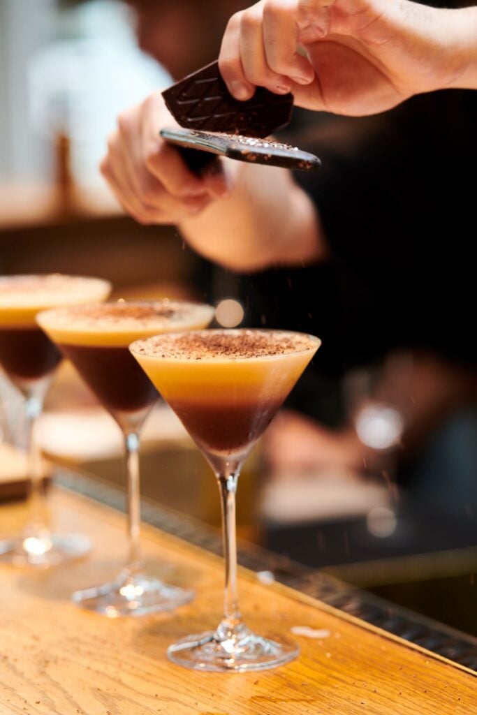 A bartender grates chocolate over three layered cocktails in elegant glasses, showcasing a rich brown and creamy top layer.