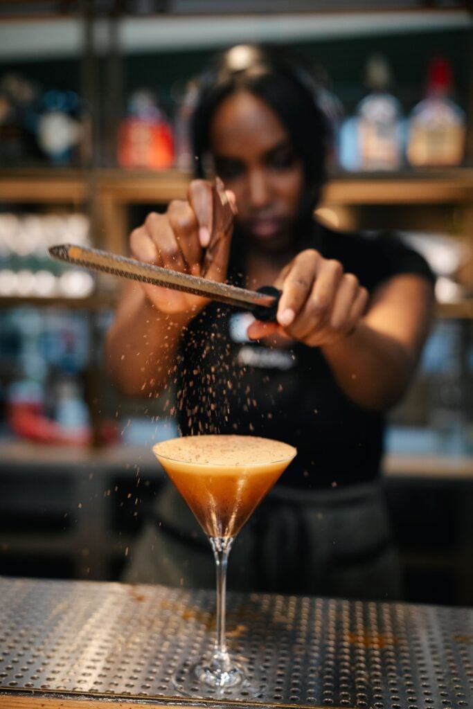 A bartender is grating chocolate over a cocktail in a martini glass, with a blurred background of bottles on shelves.