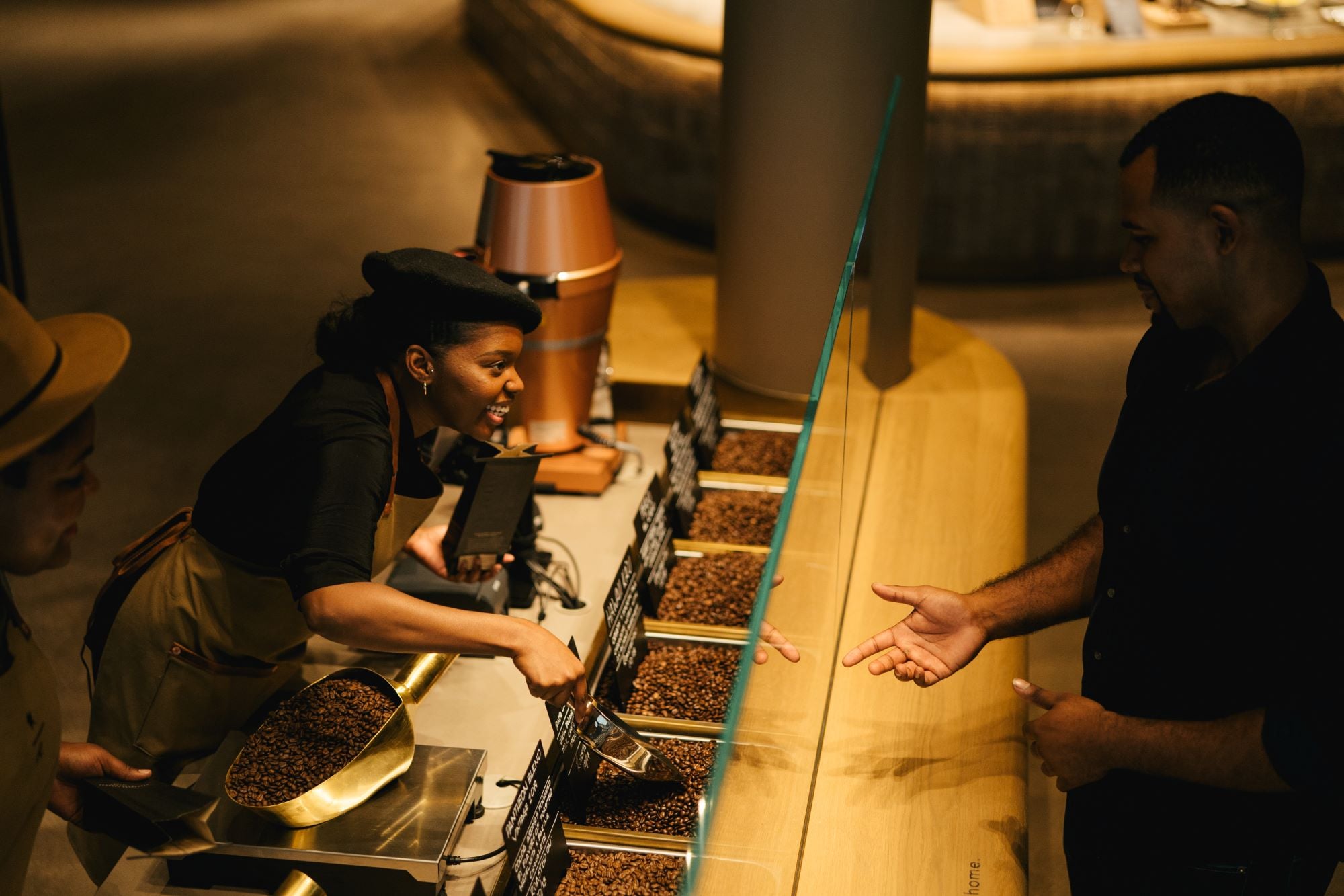 A barista in a black shirt and beret smiles while scooping coffee beans from a container. A customer reaches out with their hand.