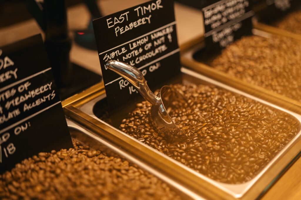 Coffee beans are displayed in a metal container with a scoop resting on top. A sign indicates "East Timor Peaberry," describing flavor notes of lemon, mint, and dark chocolate.