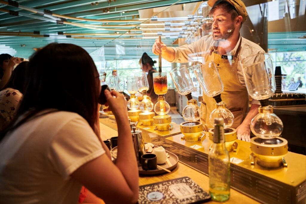 A barista prepares coffee using a siphon brewing method, with glass apparatus and glowing lights. Customers watch intently.