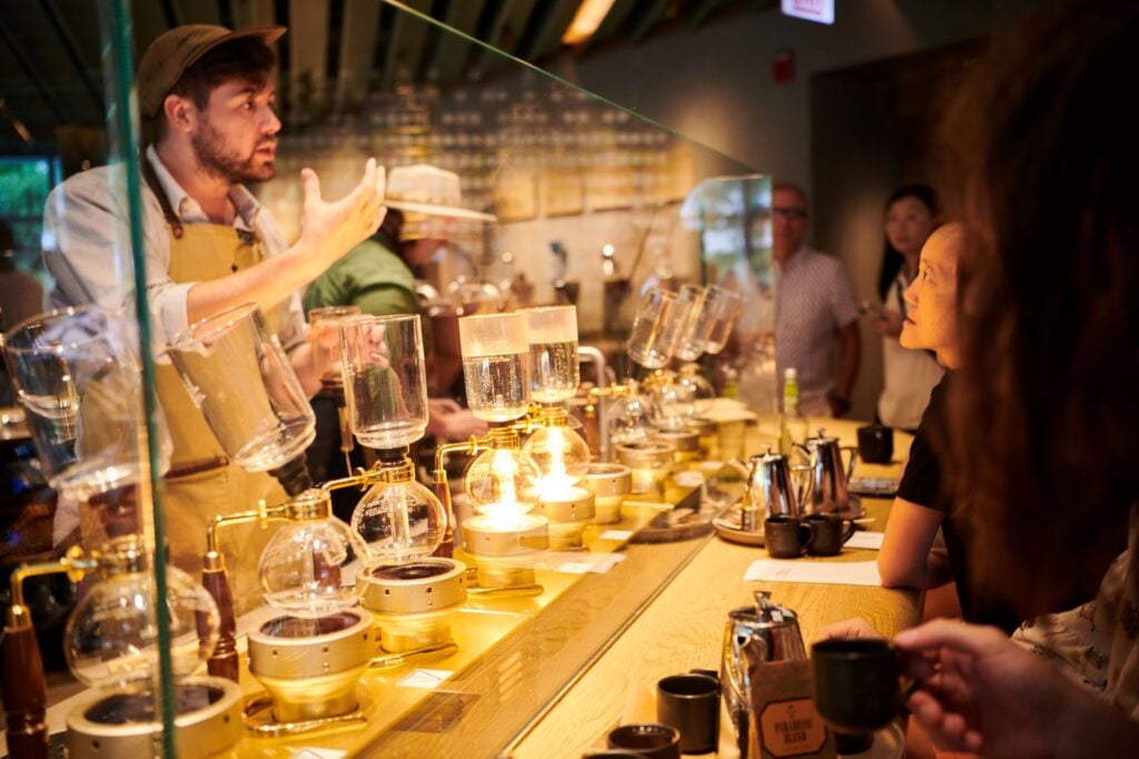 A barista demonstrates coffee brewing techniques using a siphon setup, with glass vessels and a warm ambiance. Observers watch intently.