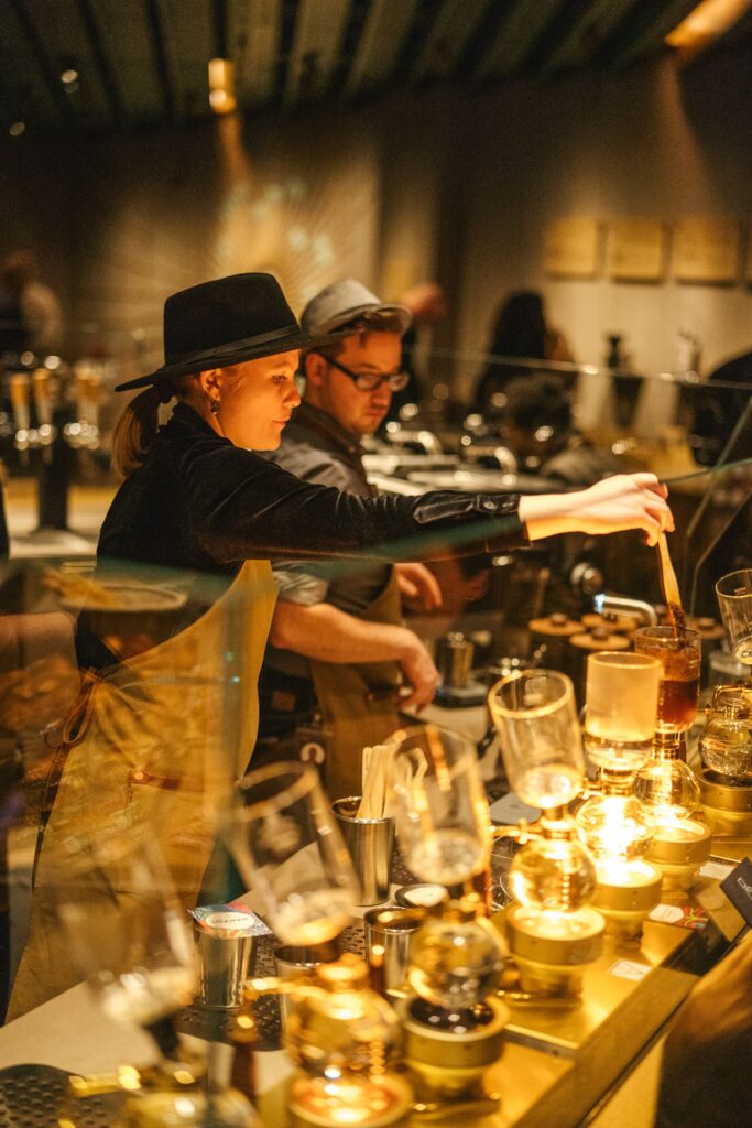 A barista in a black hat and apron prepares a drink at a stylish coffee bar, with various brewing equipment and glowing lights.