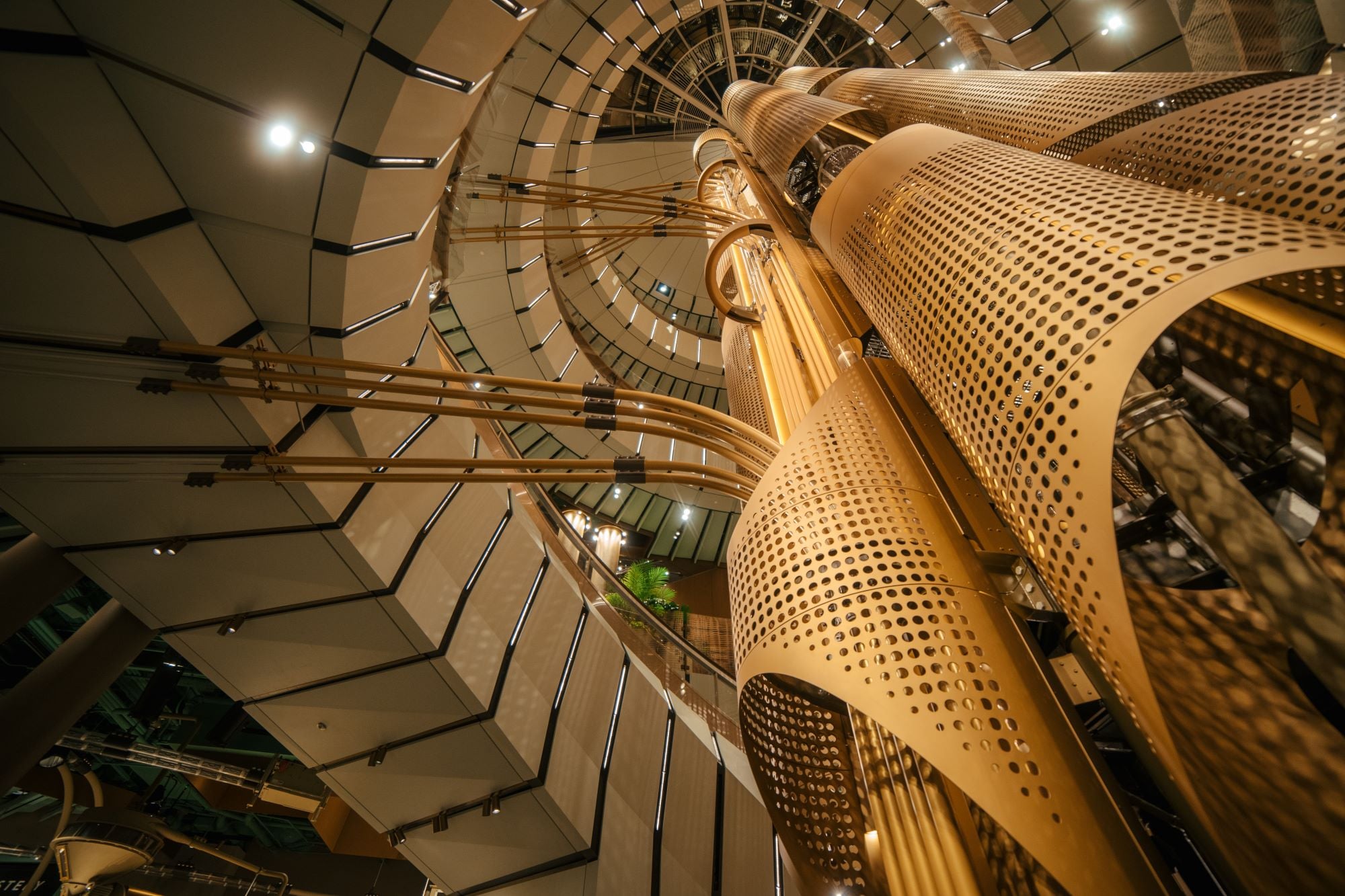 A view from below of a modern architectural structure featuring large, golden metallic columns and curved, perforated panels. The design includes sleek lines and a spiral staircase, with soft lighting illuminating the space.