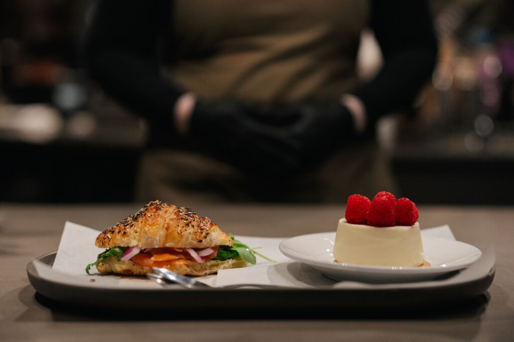 A croissant sandwich with sesame seeds, filled with greens, smoked salmon, and onions, alongside a panna cotta topped with raspberries.