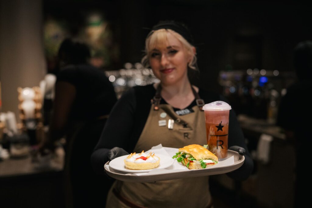 A server holds a tray with a sandwich, a dessert topped with cream and a strawberry, and a colorful drink. The background is blurred.
