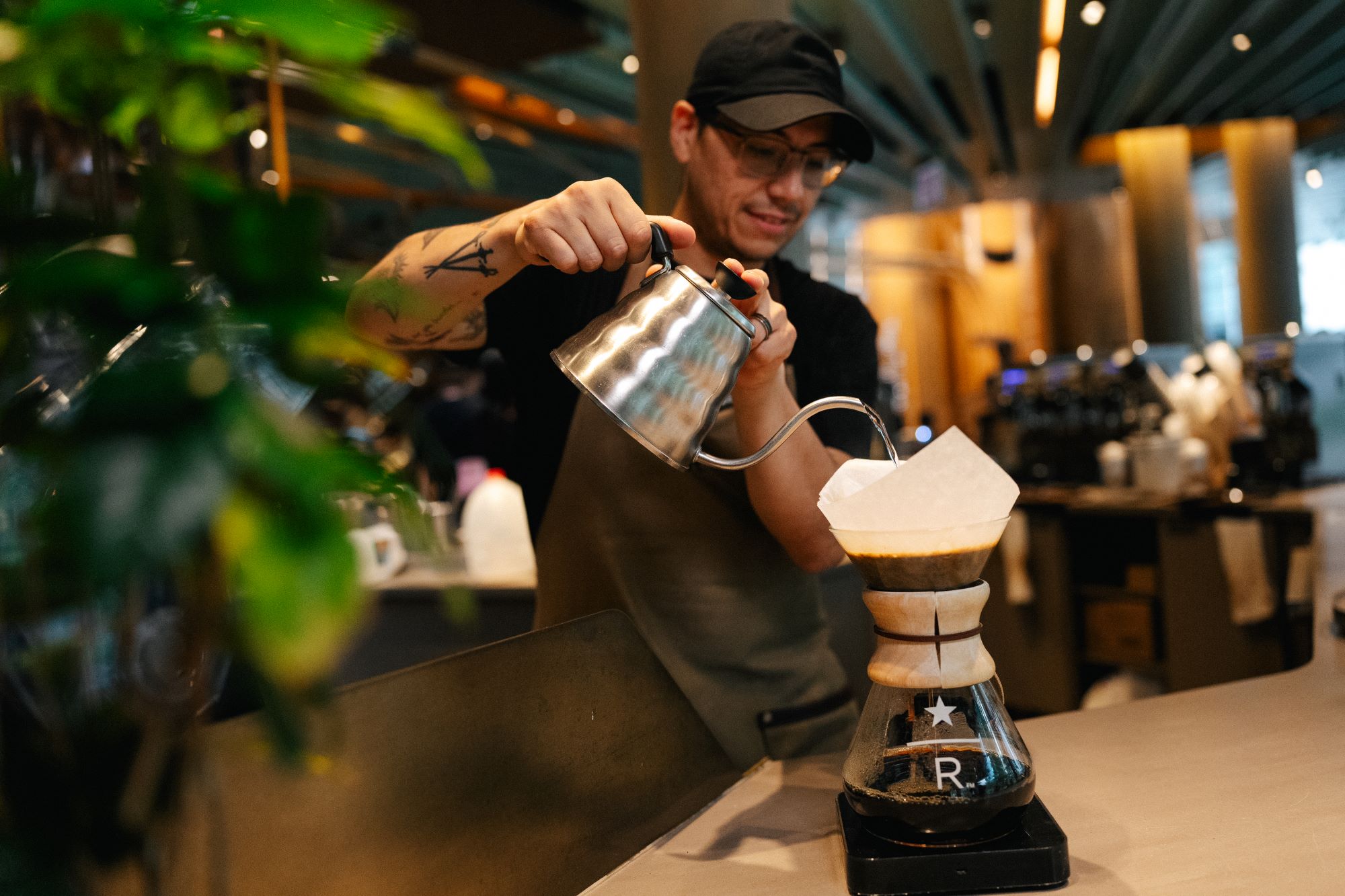 A barista is pouring hot water from a kettle into a Chemex coffee maker, which has a paper filter. The setting is a modern café.