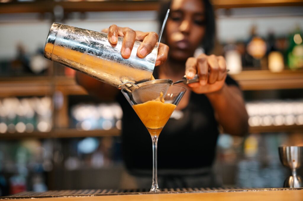 A bartender is pouring a cocktail through a strainer into a martini glass. The drink has a smooth, creamy texture and a golden color.
