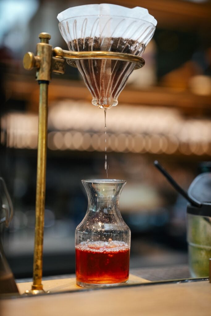 A glass coffee dripper is pouring a dark red liquid into a glass carafe. The setup features a brass stand and a paper filter filled with coffee grounds. Bokeh lights are visible in the background.