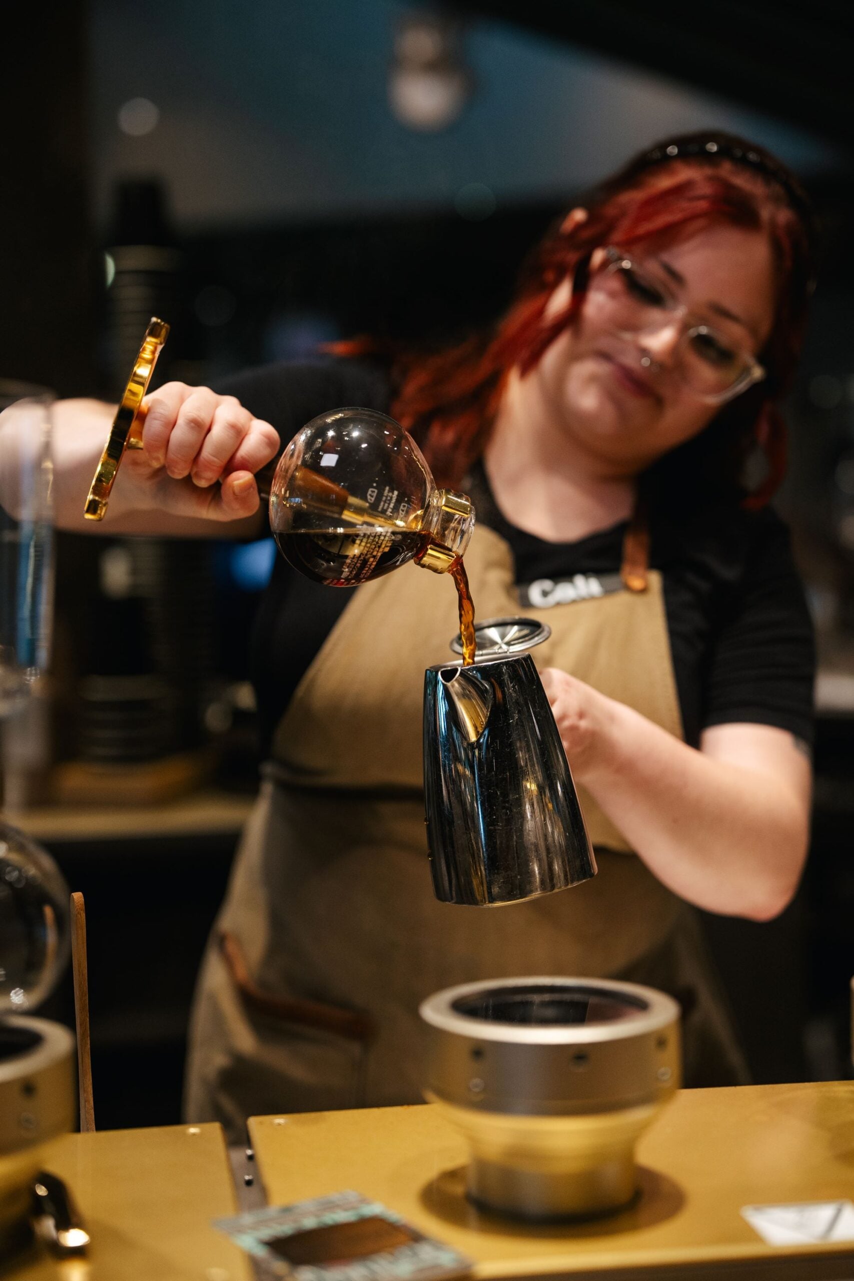 A barista with red hair pours coffee from a glass vessel into a sleek metallic kettle. The setting features coffee brewing equipment.