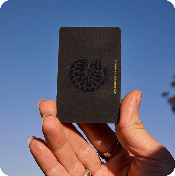 A hand holds a black Starbucks Rewards card against a clear blue sky. The card features a logo and the text "STARBUCKS REWARDS" in gold.