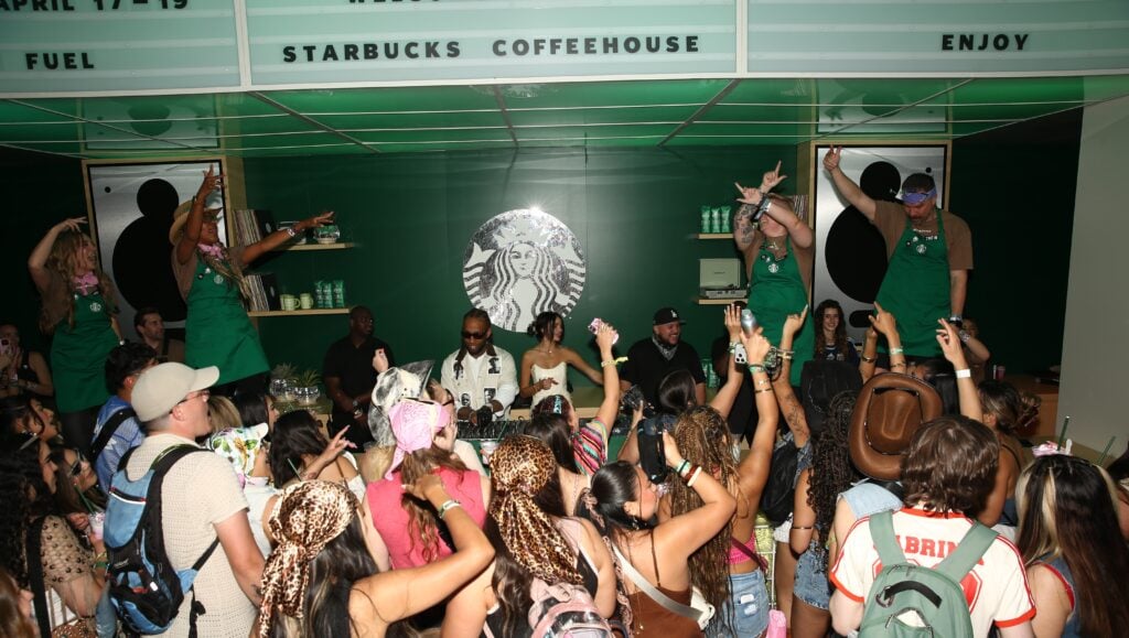 A lively crowd is gathered in a Starbucks coffeehouse, with people dancing and raising their hands. Several individuals are behind a counter, engaging with the audience. The decor features green tones and a prominent Starbucks logo.