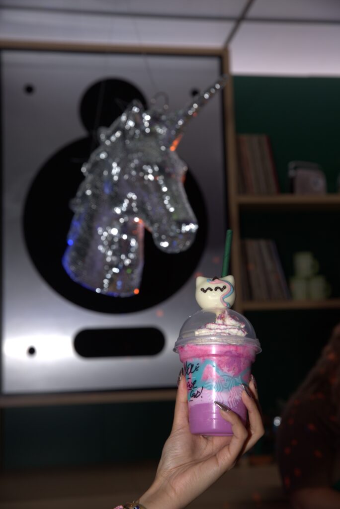 A colorful pink drink topped with whipped cream and a unicorn-shaped cookie is held up against a sparkling unicorn decoration in the background.