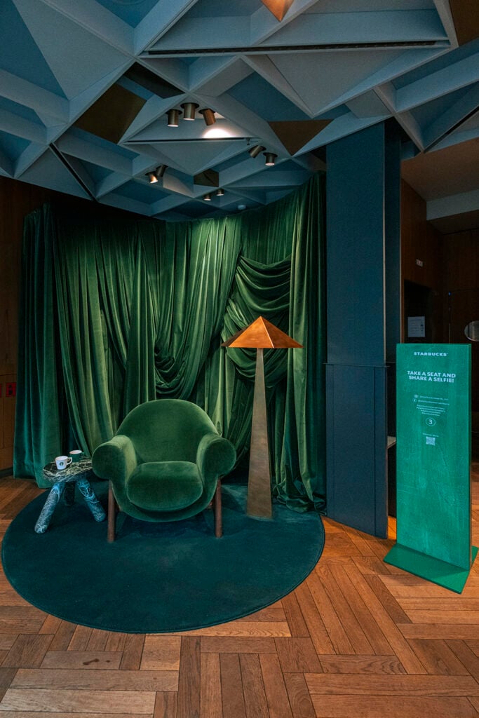 A cozy green velvet chair is positioned against a backdrop of flowing green drapes. Nearby, a geometric lamp with a copper top and a small table with a cup sit on a circular green rug, all set on a wooden floor. A sign invites guests to take a selfie.