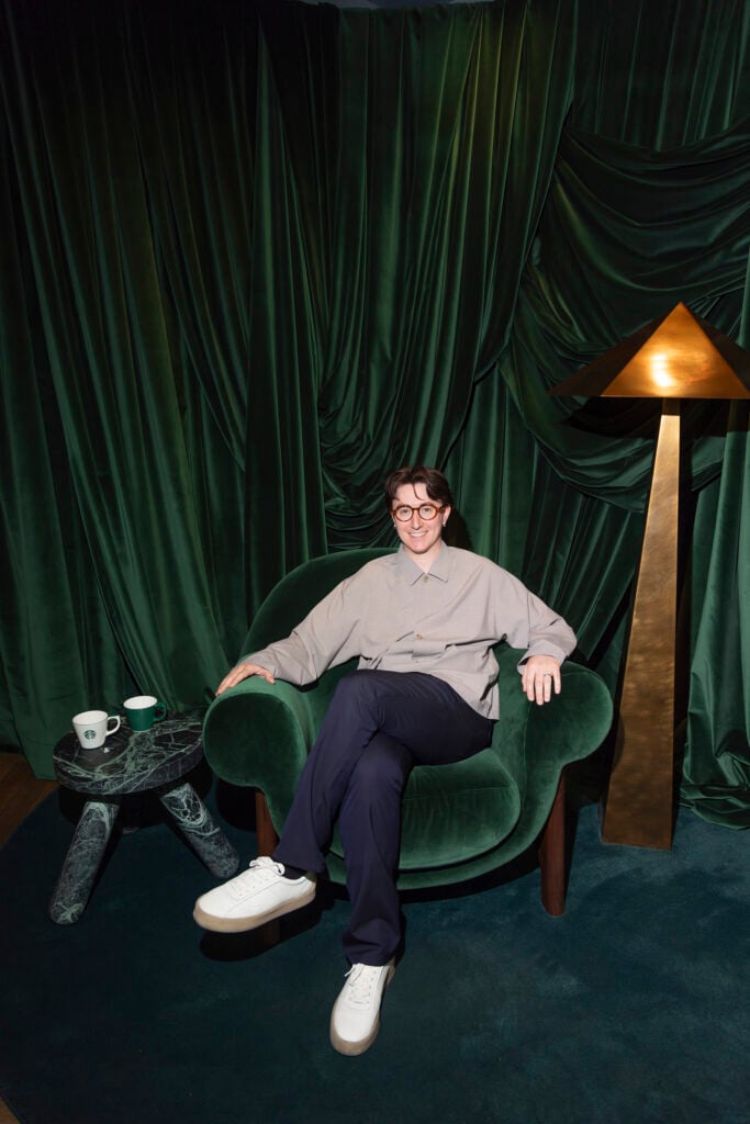 A person sits comfortably in a green velvet chair, wearing a light gray shirt and dark pants. Nearby is a marble side table with two cups, one green and one white. A gold lamp stands beside the chair, and the backdrop features rich green drapery.