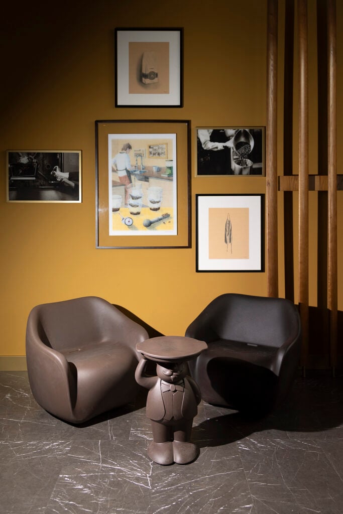 A cozy seating area features two brown, modern chairs and a whimsical brown figure resembling a waiter holding a tray. The wall is adorned with various framed artworks, including illustrations and photographs, against a warm yellow backdrop.