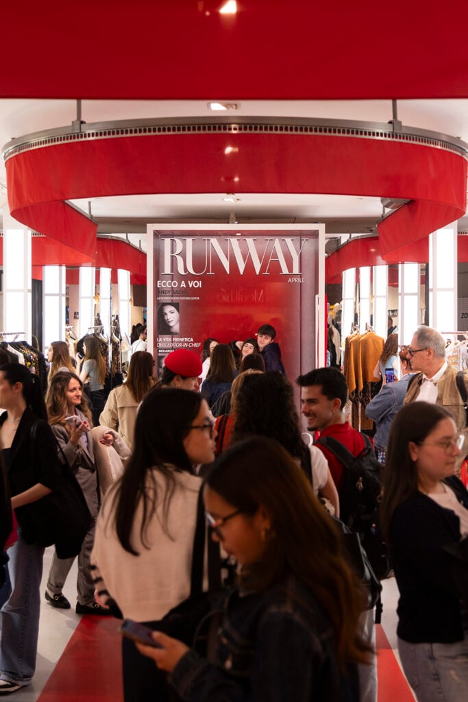 A vibrant fashion event scene with a crowd of people browsing clothing racks. A large poster titled "RUNWAY" is prominently displayed. Red accents dominate the decor.
