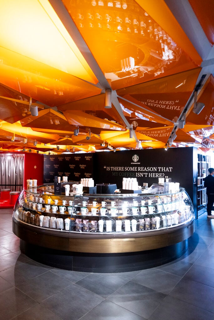 A modern coffee shop interior features a circular display counter filled with various coffee mugs and bags. The ceiling has vibrant orange panels reflecting light, and a black wall displays a quote.