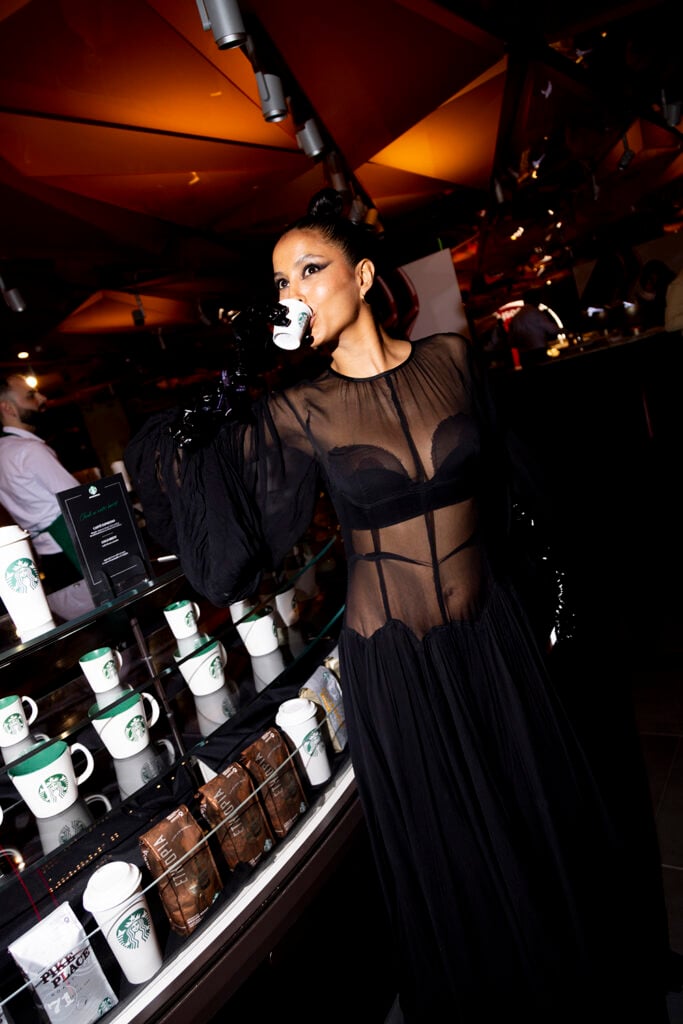 A woman in a sheer black dress poses with a coffee cup in hand. Behind her is a display of Starbucks mugs and coffee bags. The setting has warm lighting and a modern aesthetic.
