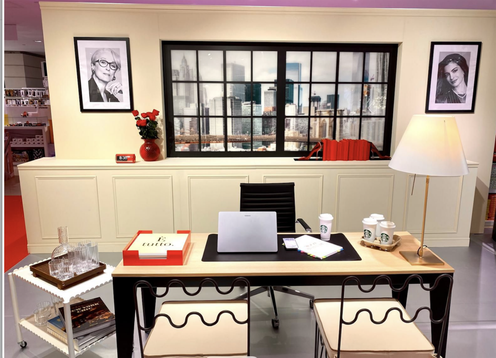 A stylish office setup featuring a modern desk with a laptop, coffee cups, and decorative items. The backdrop includes a cityscape window and framed portraits. A lamp and a side table with glassware add to the ambiance.