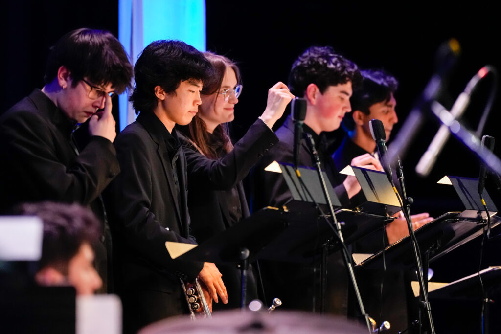 A group of young musicians in black attire perform on stage, focused on their instruments and sheet music, with microphones in front.