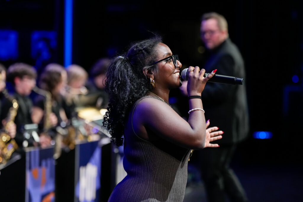 A singer with curly hair and glasses performs on stage, holding a microphone. She wears a fitted dress and gestures expressively. In the background, musicians are visible, engaged in playing instruments.