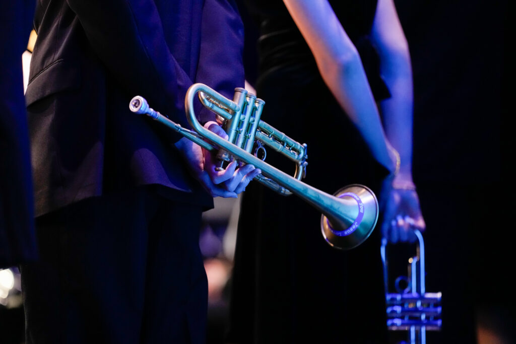 A person holds a trumpet behind their back, while another individual stands nearby, partially visible. The scene is illuminated with blue lighting.