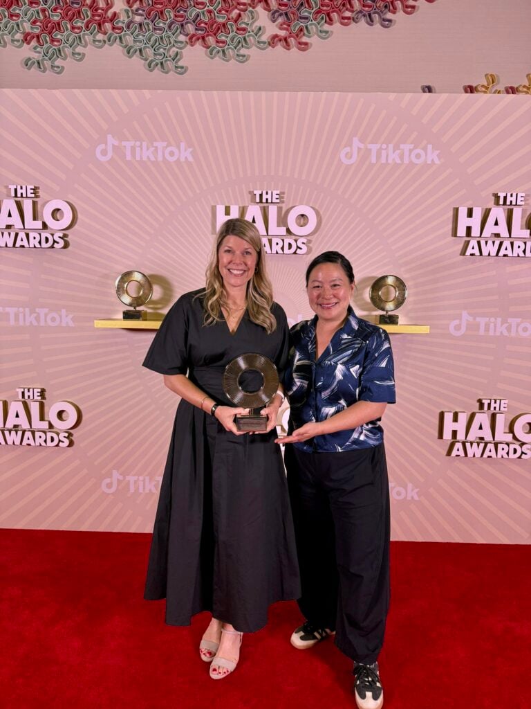 Two women stand on a red carpet in front of a backdrop featuring "The Halo Awards" and TikTok branding. One woman holds a trophy, while both are smiling. The backdrop has a decorative pattern above them.