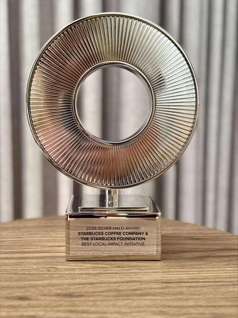 A silver award trophy featuring a circular design with radial lines, mounted on a square base. The base is engraved with text indicating it is the "2026 Silver Halo Award" for "Best Local-Impact Initiative" by Starbucks Coffee Company and The Starbucks Foundation. The background shows a soft, neutral-colored curtain.