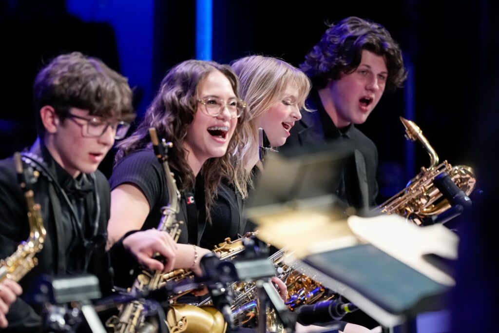 A group of young musicians joyfully perform together, playing saxophones. They are dressed in black and appear engaged in the music.