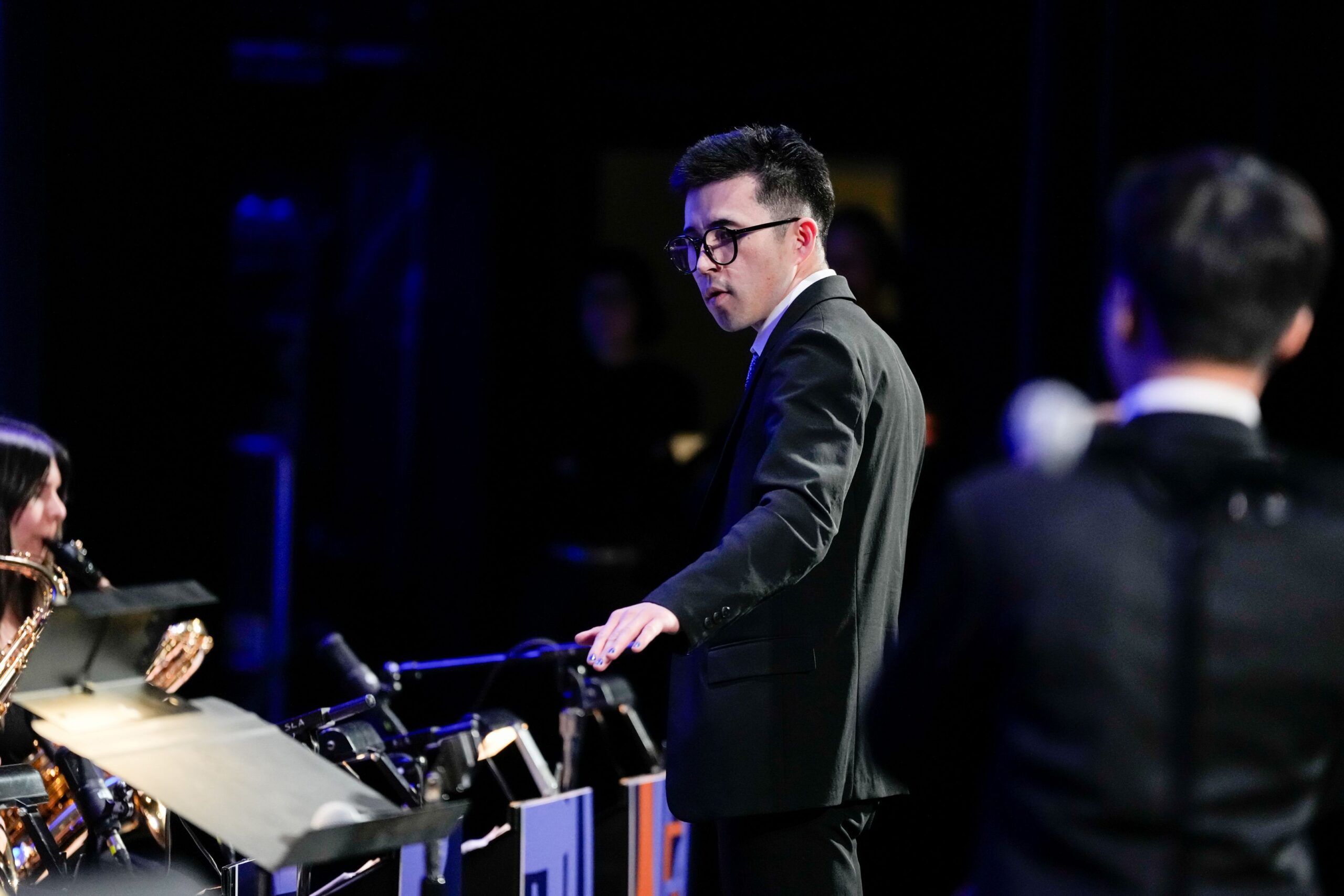 A man in a suit stands in a performance setting, gesturing with one hand. He has glasses and short hair, with a focused expression. The background is dimly lit, highlighting the stage elements.