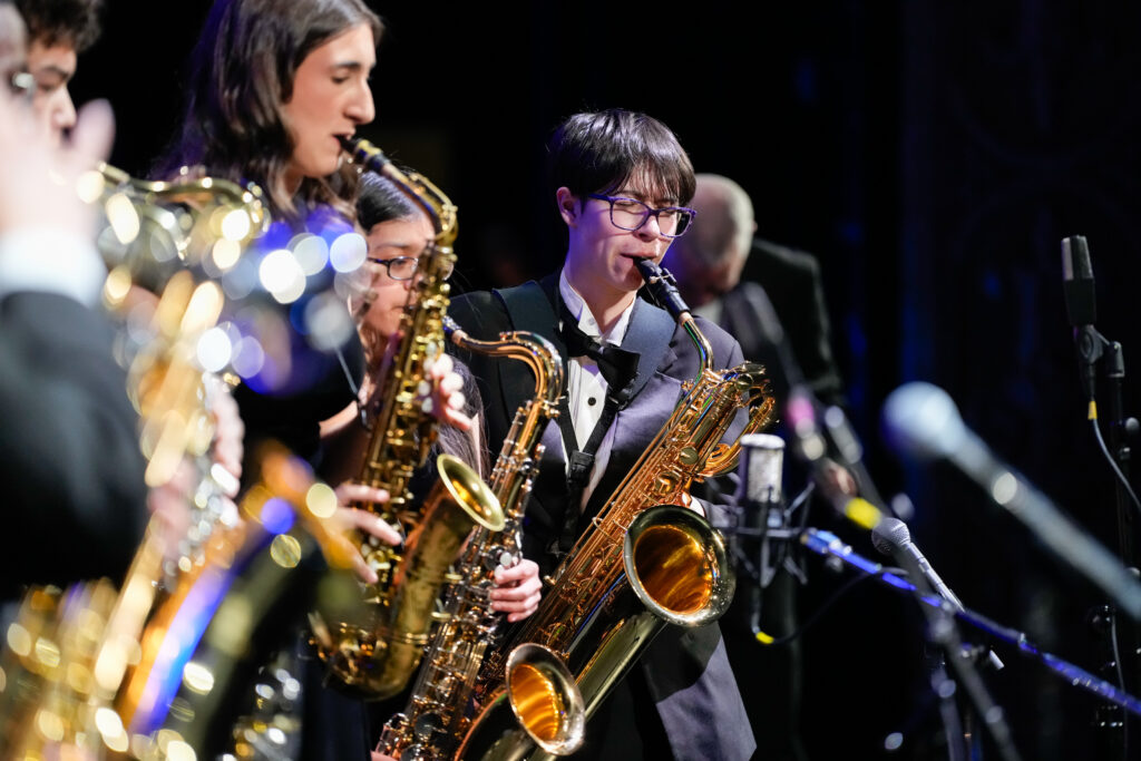 A group of musicians plays saxophones on stage, dressed in formal attire. The focus is on a performer with short hair and glasses, playing a saxophone. Other saxophonists are partially visible, creating a vibrant musical atmosphere.