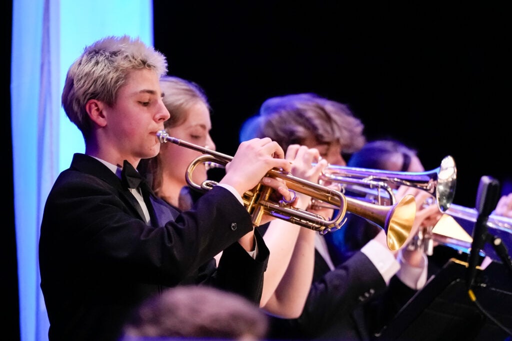 A group of young musicians in formal attire play trumpets on stage, focused on their performance. The background features soft blue lighting.