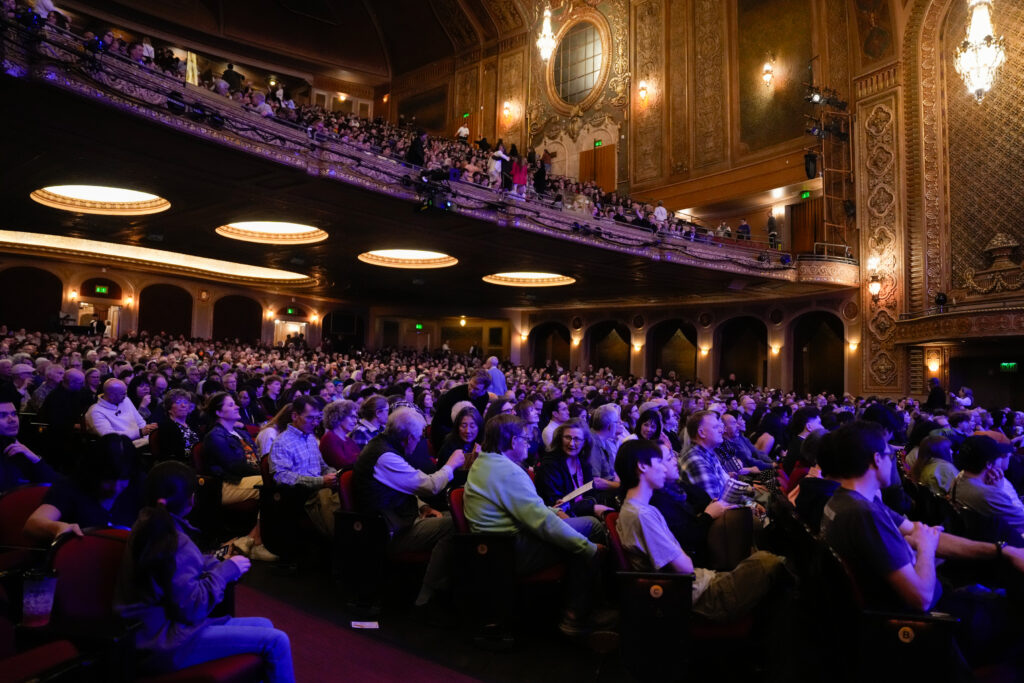 A large audience fills a beautifully ornate theater, with people seated in rows and a balcony above. The atmosphere is lively and engaged.
