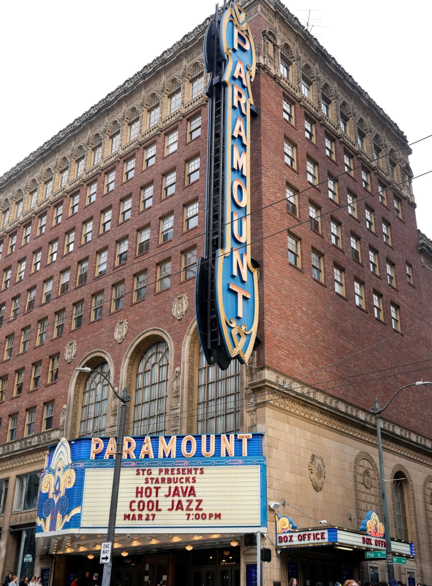 The Paramount Theatre features a large, illuminated marquee displaying "STG PRESENTS STARBUCKS HOT JAVA COOL JAZZ MAR 27 7:00 PM." The building showcases ornate architectural details with a brick facade and large windows. A line of people is visible in front, with some pedestrians and a cyclist nearby.