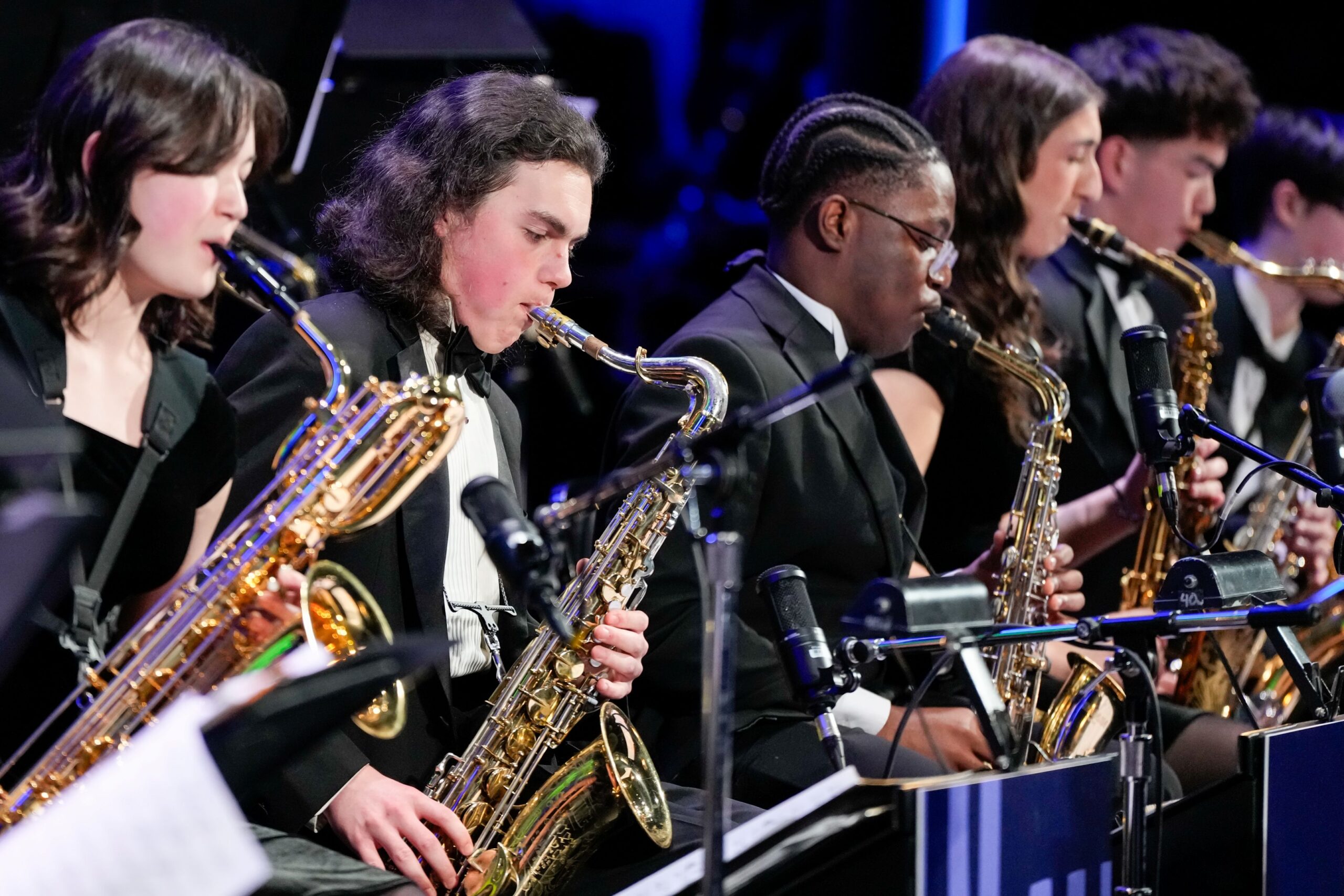 A group of musicians playing saxophones on stage, dressed in formal black attire. Microphones are positioned in front of them.