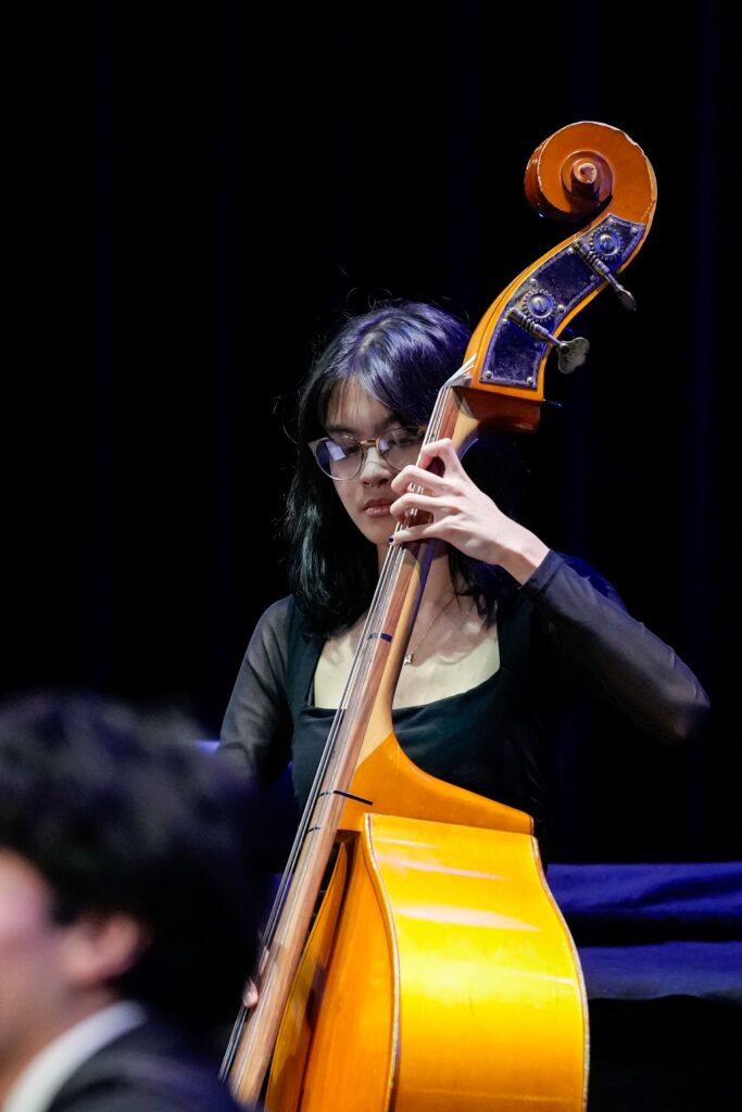 A musician plays a double bass, wearing glasses and a black outfit, focused on her performance against a dark background.