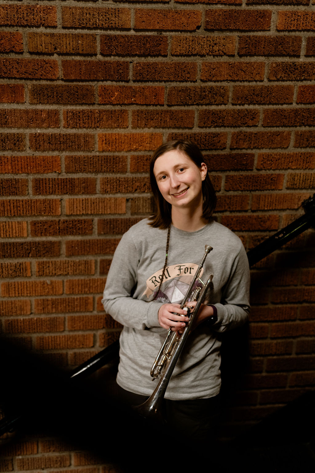 A person stands against a brick wall, holding a trumpet. They wear a gray sweatshirt with a graphic design and smile at the camera.