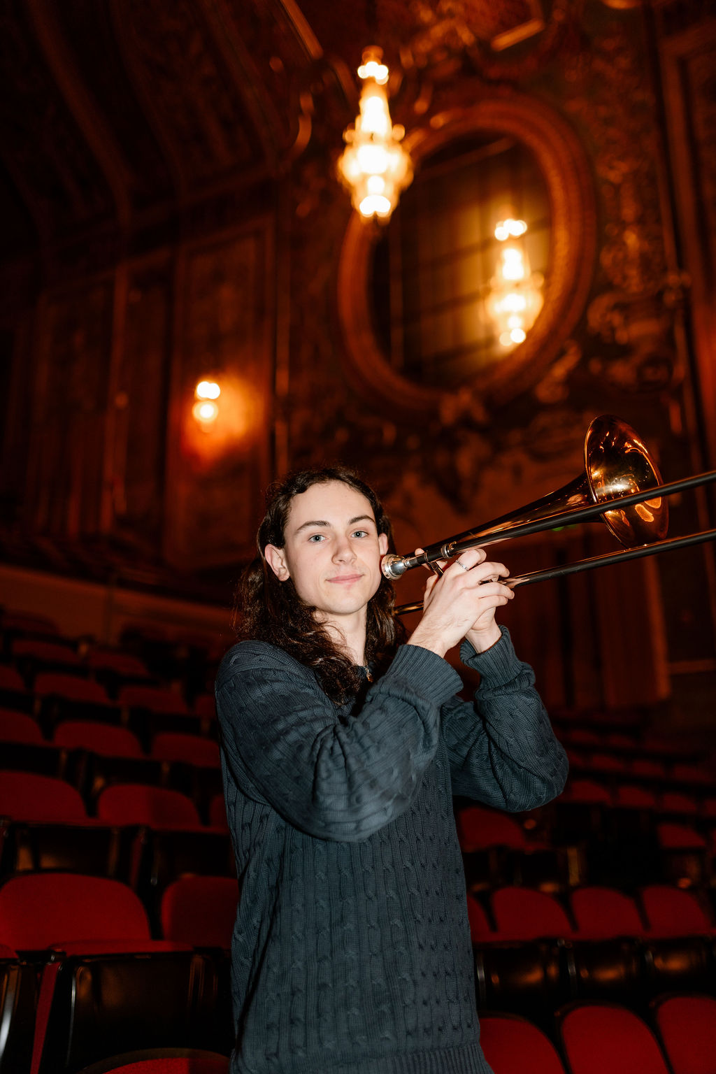 A young person with long hair holds a trombone, standing in an ornate theater with red seats and decorative lighting in the background.