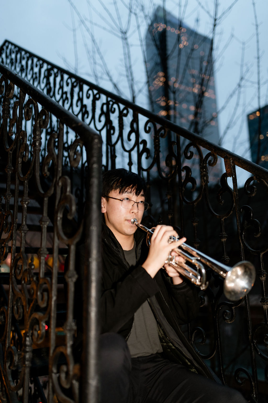A young musician plays a trumpet while seated on a staircase, surrounded by ornate iron railings. The background features a cityscape at dusk.