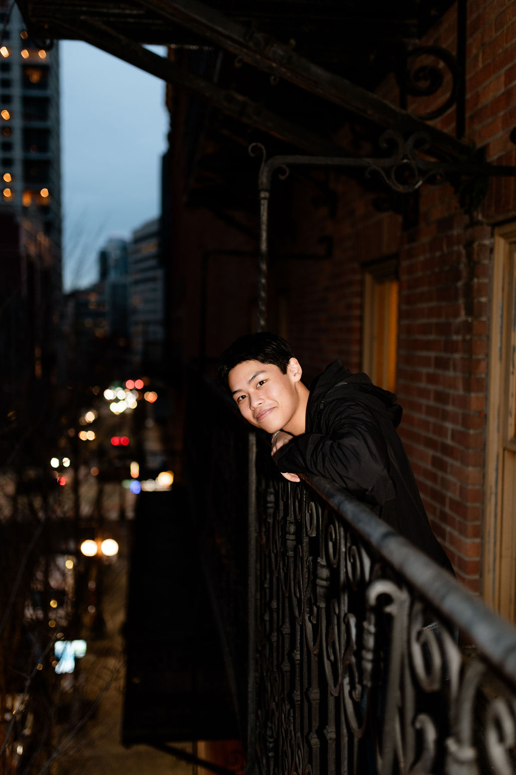 A young person leans on a wrought iron railing on a balcony, smiling. The background features a city street with blurred lights, suggesting evening.